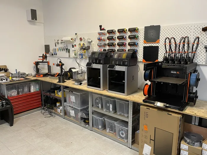 Werkstatt mit mehreren 3D-Druckern auf einer langen Werkbank, darüber eine Lochwand mit Werkzeug und Filamentspulen